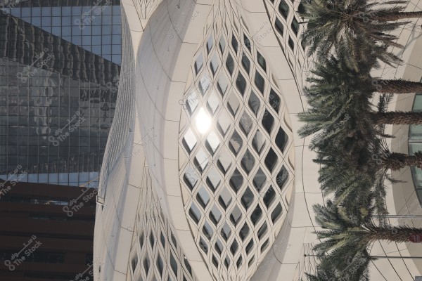 An image of a modern building facade with a unique architectural design during the daytime. The facade consists of white structures with oval lattice patterns, and light is reflecting on the glass surface. To the right, there are palm trees, and the background of the building is covered with glass that intriguingly reflects the surroundings.