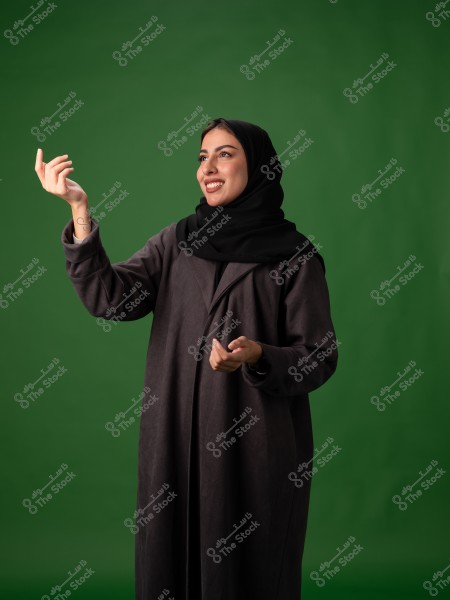 ** A portrait of a woman wearing a black abaya and black hijab, standing in front of a green background. The woman is smiling and looking upwards with an expressive hand gesture.\r\n\r\n**