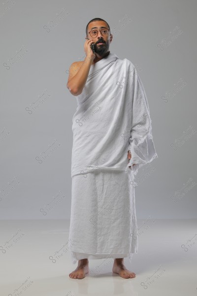 A man stands barefoot on a white floor, wearing the white garments of Ihram, consisting of an izar and rida\'. He is holding a mobile phone and speaking into it. The man has a short beard and wears glasses. The background is plain gray.