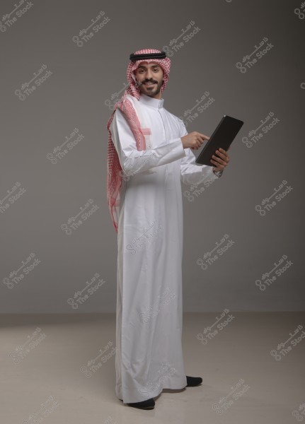 Portrait of a smiling man standing, wearing a white thobe, a red and white checkered ghutra, and a black agal, pointing at a tablet with his right hand. The photo seems to be taken in a studio with a simple gray background.\r\n\r\n###