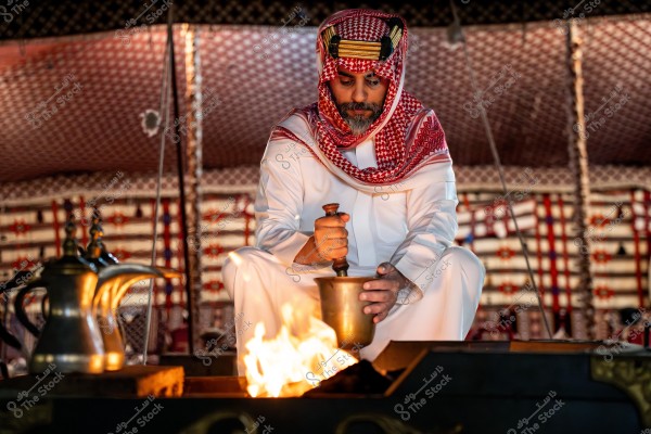 A man sitting in a traditional tent, wearing a white thobe and a red and white headscarf, likely from Saudi Arabia. He is holding a brass pestle and mortar over a burning fire. The background of the tent features traditional woven patterns.