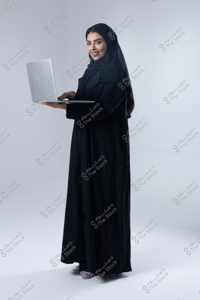 صورة لامرأة ترتدي عباءة سوداء وحجاب, وتقف بينما تحمل جهاز كمبيوتر محمول. يبدو أنها تبتسم وتنظر إلى الكاميرا. الخلفية بيضاء وخالية.