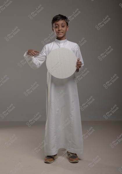 The image shows a boy wearing a long white thobe standing in a studio with a gray background. The boy is holding a large white circle and pointing at it with his other hand. The thobe looks traditional and possibly of Saudi origin. The boy is wearing brown athletic shoes.