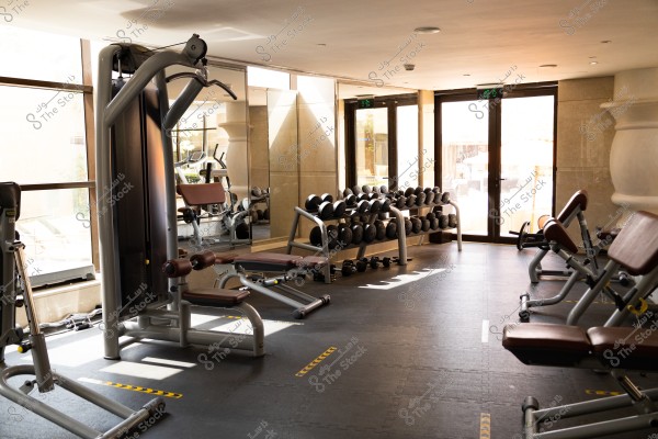 صالة رياضية حديثة ومشرقة مع معدات تمارين القوة مثل الأثقال وأجهزة متعددة التمارين. تُظهر الصورة مجموعة من الأوزان مرتبة بدقة أمام مرآة كبيرة. تتخللها إضاءة طبيعية من النوافذ الكبيرة وتنعكس على الأرضية المبطنة باللون الداكن. التفاصيل الداخلية تتضمن الأثاث الرياضي المقاوم والمعدات المتنوعة.