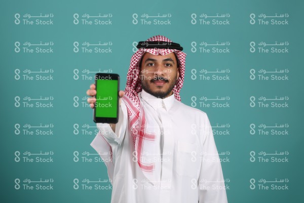 بورتريه رجل سعودي يرتدي الثوب ويقف أمام خلفية تركوازية ويظهر هاتفه