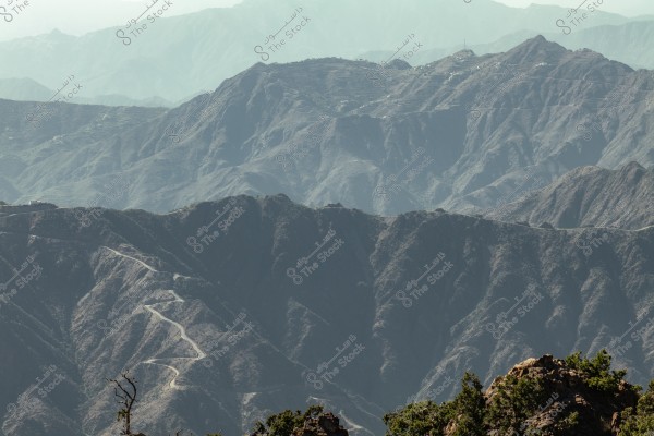A landscape view of a mountain with rugged terrain and multiple elevations. The dark gray mountains stretch across the horizon. A long, winding road runs along the mountain, traversing steep slopes, with scattered trees in the foreground.