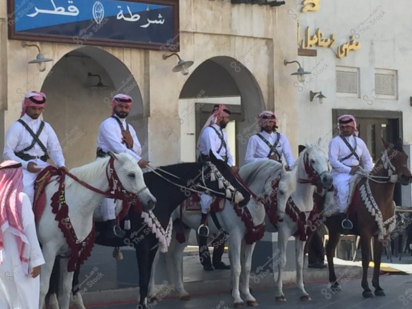 The image shows a group of men wearing traditional Qatari attire, riding horses adorned with traditional decorations. The men are dressed in white thobes and red headscarves. A sign in the background reads \"Police of Qatar,\" indicating the location might be Qatar.