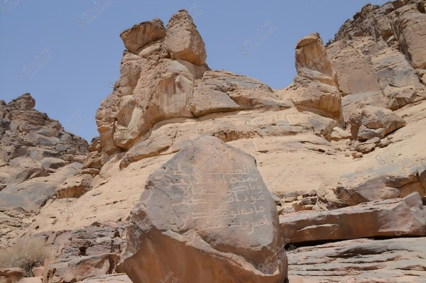 View of a large rock in a desert environment with Arabic inscriptions. The rock is surrounded by massive rock formations under a clear blue sky. The inscriptions appear historical and may date back to an ancient period.