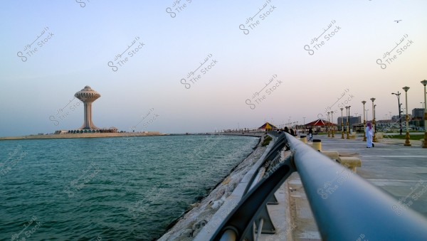 The image shows the Al Khobar Corniche in Saudi Arabia with the waters of the Arabian Gulf in the foreground. An iconic water tower is visible on the left side, surrounded by calm blue waters. A paved path extends on the right side bordered by a metal railing, with people walking along the promenade. Background buildings line the shore under a clear sky during sunset.