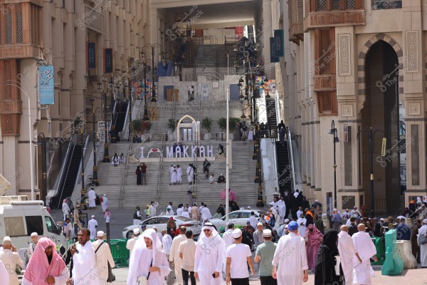 The image shows a busy scene in an area of Mecca, with numerous people wearing traditional clothing such as white thobes and ghutras. The surrounding buildings feature traditional Arabic architectural design. Large stairs are centered in the image with a sign saying \"I ♥ Makkah\". The location includes several escalators and ornate columns, and the buildings are multi-storied. The area is bustling with people walking and vehicles passing through.