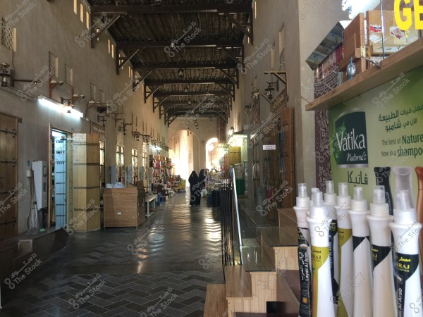 An image of a corridor in a traditional indoor market. On the left side, there are a series of shops or stalls with closed wooden doors. The floor is tiled with an interlocking pattern. On the right, shelves display beauty and hair care products from well-known brands. In the background, two people can be seen wearing traditional abayas. The ceiling is crafted from wood with some ventilation openings.
