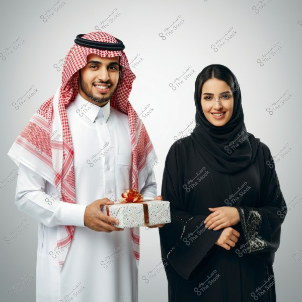 An image of a smiling man and woman. The man is wearing traditional Saudi attire with a red and white checkered shemagh and is holding a gift wrapped with a brown ribbon. The woman is wearing a black abaya with intricate embroidery and a black hijab.