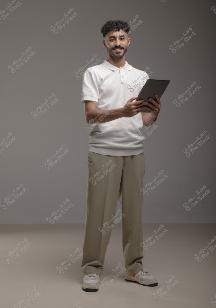 A portrait of a man standing and smiling while holding a tablet. He is wearing a white polo shirt and light brown pants with white sneakers. The background is gray, highlighting his modern and casual appearance.