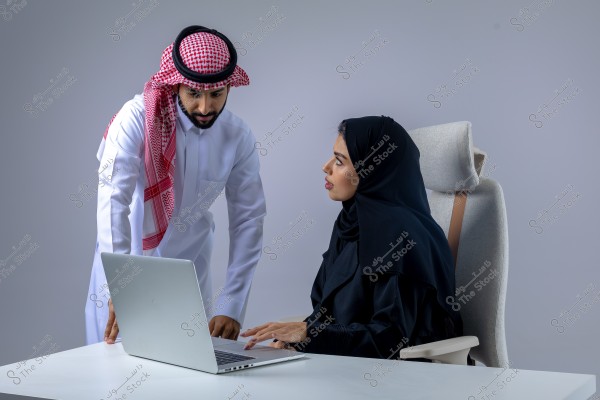 An image showing a man and a woman in an office. The man is wearing traditional Saudi attire, a white thobe and red shemagh, and appears to be speaking to the woman seated. The woman is wearing a black abaya and hijab, sitting in front of a laptop on a white desk. The setting reflects a modern work environment.