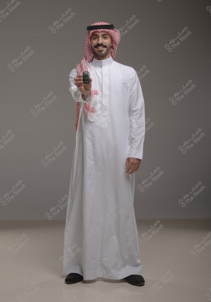 A portrait of a man wearing traditional Saudi attire, a white thobe and a red checkered headscarf (ghutra) with a black agal. He stands against a grey background, smiling and looking at the camera while holding a car key in his extended right hand.
