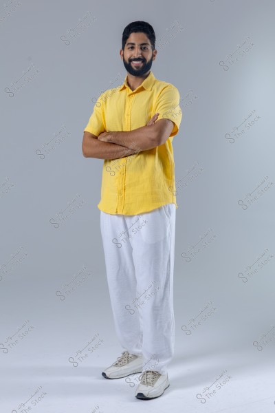 A portrait of a man wearing a yellow shirt, white pants, and white sneakers, standing and smiling with his arms crossed. The background is a simple gray, highlighting the individual in the image.