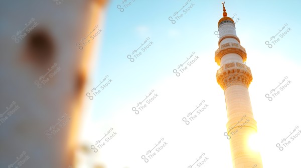 Image of a tall white mosque minaret under a clear blue sky. The sun is positioned behind the minaret, creating a soft glow around it.