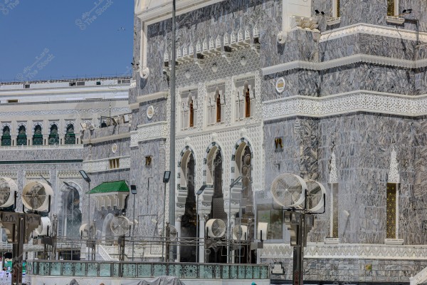 Image of a historical building with an ornate facade made of gray and white marble, featuring arched windows adorned with Islamic art. Large fans are placed in the foreground for cooling the atmosphere. The weather appears sunny and clear, accentuating the authentic Islamic architectural style.