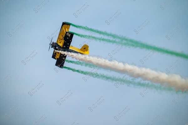A vintage biplane flying in the blue sky, emitting colored smoke in white and green. The wings are painted black and yellow with white star symbols on the lower wing.