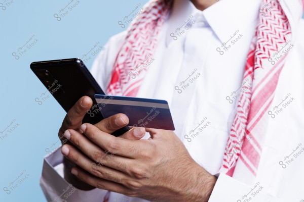 A man wearing traditional Saudi attire, a white thobe, and a red and white shemagh, holding a smartphone and a credit card. The image focuses on his hands as he uses the phone and interacts with the card. The background is blue.