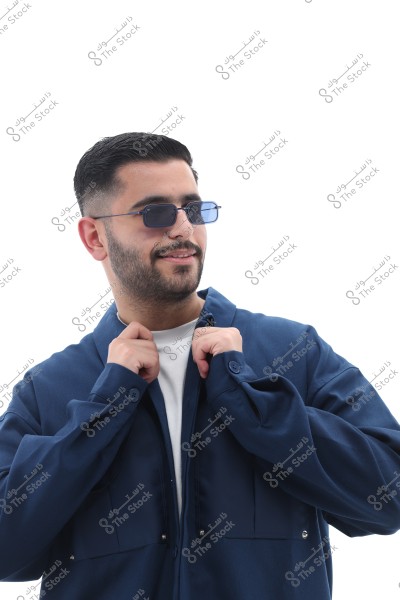 A portrait of a man wearing a blue shirt and sunglasses with blue lenses, appearing to adjust the collar of the shirt with his fingertips. The background is plain white, making the man the focal point of the image. His facial expression appears happy and optimistic.