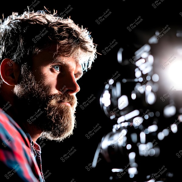 A portrait of a man with a thick beard and mustache, looking to the side. He is wearing a plaid shirt. Camera equipment is visible in the background with a bright light shining. His facial features are highlighted against the dark background due to the lighting.\r\n\r\n-
