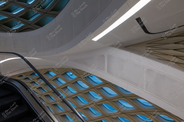 An image of an interior ceiling with a modern architectural design. The ceiling has a complex geometric structure with slanted lattice panels and blue shadows, allowing natural light to enter through the windows. In the foreground, part of an escalator handrail is visible, suggesting an indoor setting of an architectural structure like a museum or shopping center.