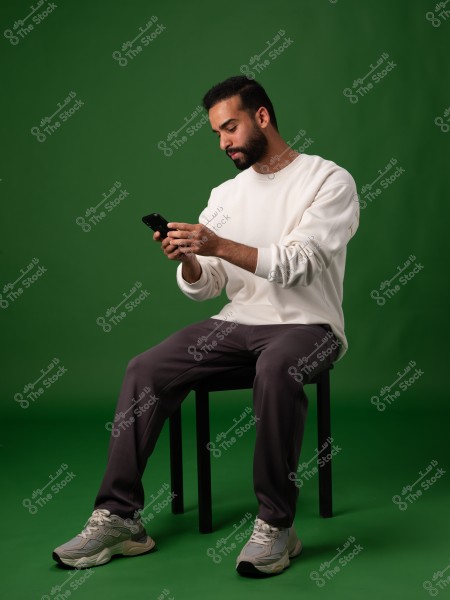 Image of a man sitting on a chair against a green background. He is wearing a white long-sleeved shirt and dark-colored sweatpants with athletic shoes. He appears focused on the phone he is holding in his hands.