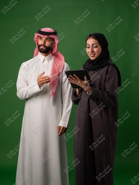 رجل يرتدي الثوب السعودي التقليدي يقف بجانب امرأة ترتدي العباءة وتحمل جهاز لوحي, على خلفية خضراء.