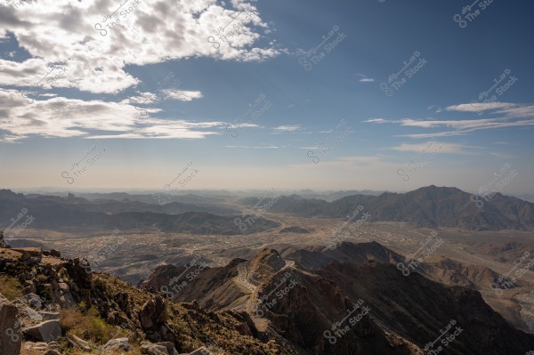 A panoramic view of towering mountains under a blue sky with scattered clouds. A winding road traverses through the mountains, reaching into the horizon, surrounded by rugged terrain and valleys. Beautiful and enchanting landscapes extend infinitely into the background.