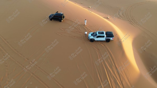 Aerial view of a vast desert under the sunset. The image features two vehicles, one black and the other white, parked on the sand. Two men wearing traditional white clothing stand near the vehicles. Tire tracks are visible on the soft sand, which reflects the gentle sunlight.