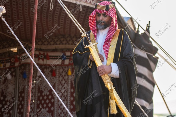 A portrait of a man standing while wearing traditional Saudi attire. The man is dressed in a white thobe with a black bisht adorned with gold trim, and a red shemagh with a black agal on his head. He holds a long, golden sword confidently, and behind him, there\'s a tent decorated with traditional patterns and various colored tassels.