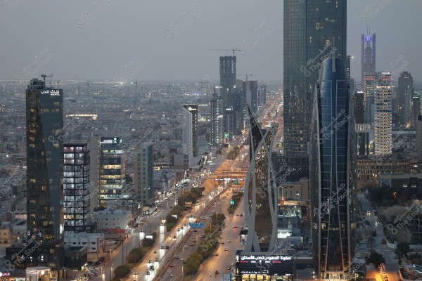 An image of Riyadh, Saudi Arabia, showing a high-altitude view of the city at dusk. The modern skyscrapers and tall buildings are illuminated with bustling streets filled with vehicles and lights. Notable buildings and modern architecture define the urban skyline of the city.