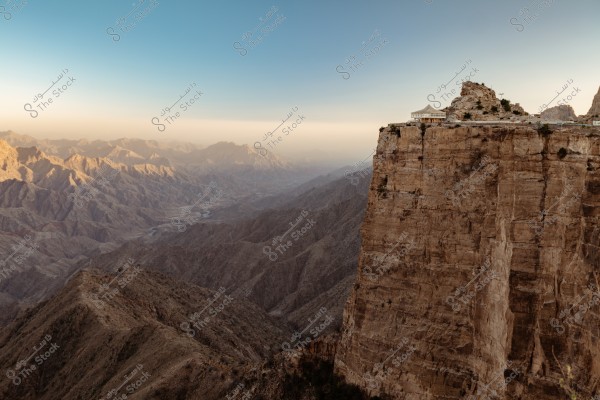 A stunning mountainous landscape is depicted in the image, featuring rocky mountains that stretch across the horizon under a clear blue sky. On the right side of the image, a high rocky cliff with a small structure, possibly a kiosk or viewing platform, is visible.