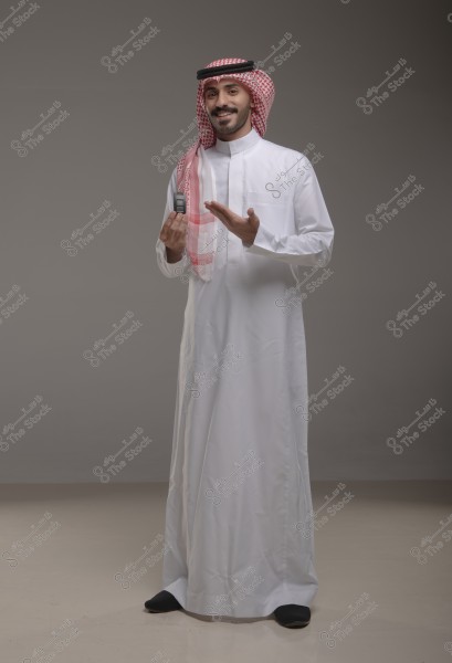 صورة لرجل يرتدي ثوباً تقليدياً أبيض وكوفيه حمراء وبيضاء وعقال أسود, قد يكون من المملكة العربية السعودية. يقف مبتسماً ويحمل جهازاً صغيراً في يده اليسرى, بينما يديه اليمنى مرفوعة بإيماءة شرح.