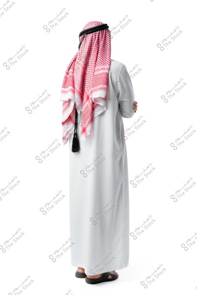 An image of a man wearing a traditional white thobe and a red checked keffiyeh with a black agal, standing with his back to the camera. The person is wearing black sandals on a white background.