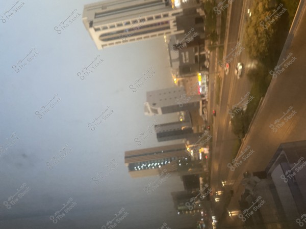 A nighttime cityscape featuring numerous high-rise buildings illuminated by yellow glowing lights. The image shows a busy road with vehicles, surrounded by commercial buildings and small landscaped areas. The sky is dark with dim lighting from the city.