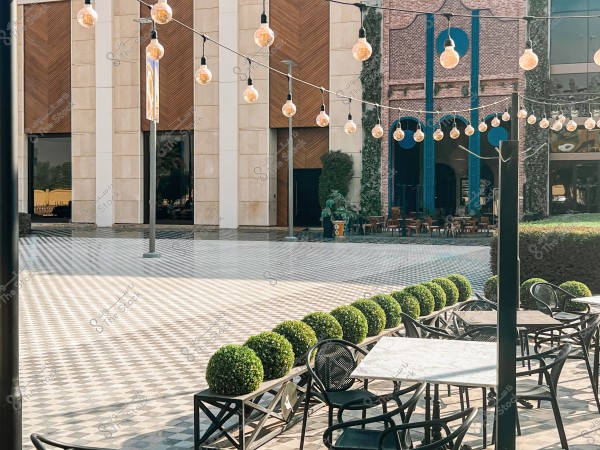 The image shows an outdoor courtyard with a checkered tile floor in light and dark colors. A line of black metal tables and chairs is positioned near some neatly trimmed shrubs. The courtyard is decorated with string lights hanging above. The background features elegant architectural buildings with brick designs and blue-painted accents.