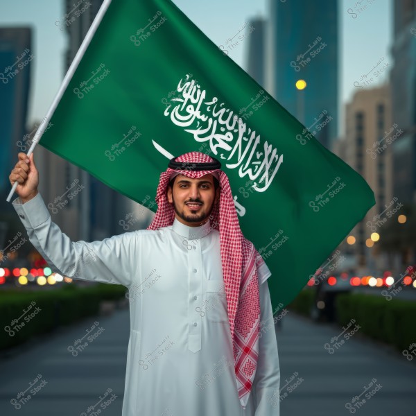 A portrait of a man wearing traditional Saudi attire, consisting of a white thobe, a red and white shemagh, and a black agal. He is smiling and holding the green Saudi Arabian flag. Urban background with city lights can be seen in the evening.