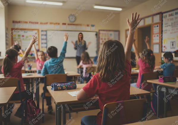 The image shows a classroom with children sitting at desks, raising their hands to answer questions from a teacher standing in front of a whiteboard. The whiteboard behind the teacher has some writing on it. The walls are decorated with educational posters, and the desks have school supplies like pencil cases.