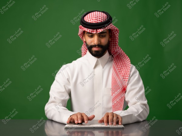 رجل يرتدي الثوب السعودي والشماغ يجلس أمام لوحة مفاتيح على خلفية خضراء.
