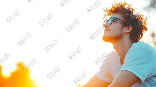 An image of a man wearing a white shirt and sunglasses, sitting outdoors with sunlight reflecting on his hair and face. He appears to be enjoying the sunny weather, with a brightly lit background that adds a warm atmosphere to the photo.