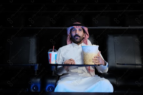 شاب سعودي يرتدي الزي السعودي التقليدي  يجلس في صالة السينما ويتفاعل أثناء مشاهدة الفيلم، رجل سعودي يرتدي الثوب والشماغ يجلس في صالة السينما ويبدو مندهشًا أثناء مشاهدة الفيلم ويظهر أمامه الفشار وأحد المشروبات على الطاولة