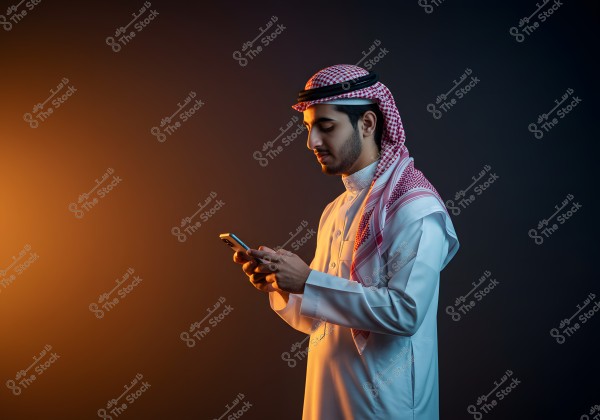 A portrait of a man wearing traditional Saudi attire, including a thobe and ghutra. He stands against a dark background, holding a phone in his hands, with warm lighting illuminating his face.
