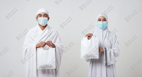 The image shows a man and a woman wearing traditional white Ihram clothing, which is specific attire for Umrah or Hajj. Both are wearing blue medical face masks. The man holds a white bag in his hands, as does the woman, who is also wearing a hijab. The background is plain white.