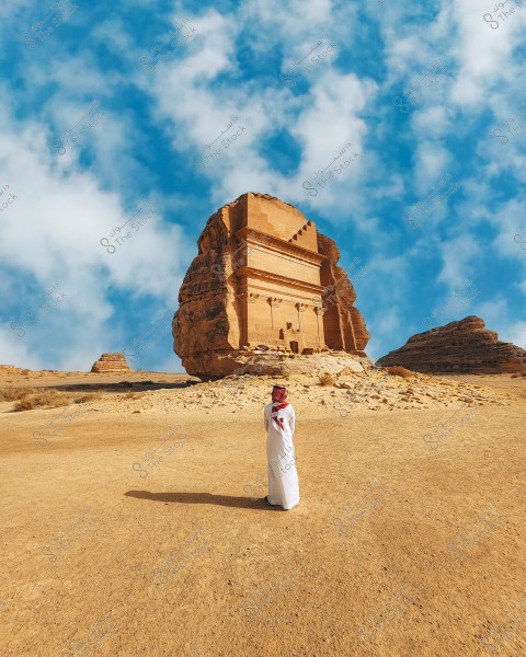 A person stands in front of a stunning rock formation in Al-Ula, Saudi Arabia. They are wearing a white thobe and a red ghutra, reflecting traditional Saudi attire. In the background, the massive and intricately carved rock structure is visible, with a blue sky and scattered clouds.