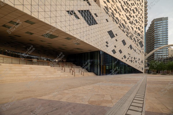 An image of a modern building with an innovative architectural design featuring a slanted facade with glass squares. The building is adjacent to a wide open plaza with paved flooring and small trees. In the background, there are other tall buildings, indicating it is part of a modern urban area.
