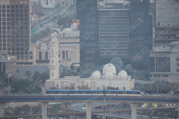 An image depicting an urban scene in Riyadh, Saudi Arabia, featuring modern buildings with glass facades and a tall minaret of a mosque. In the foreground, a blue train travels on an elevated track. Additionally, a large white mosque dome is visible in the background.