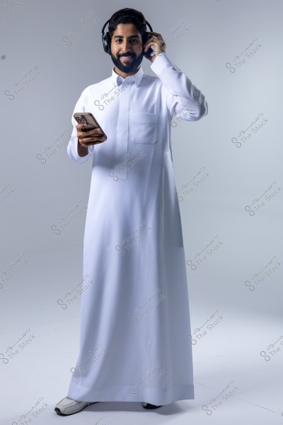 A portrait of a man wearing a traditional white thobe and black headphones. He appears happy while holding a smartphone in his hand. He stands against a white background, and the clothing suggests it may be from the Gulf region.