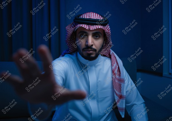 The image shows a man wearing traditional Gulf attire, with a white thobe and a red and white shemagh secured with a black agal. He appears to be sitting in a room with blue lighting, extending his hand towards the camera.